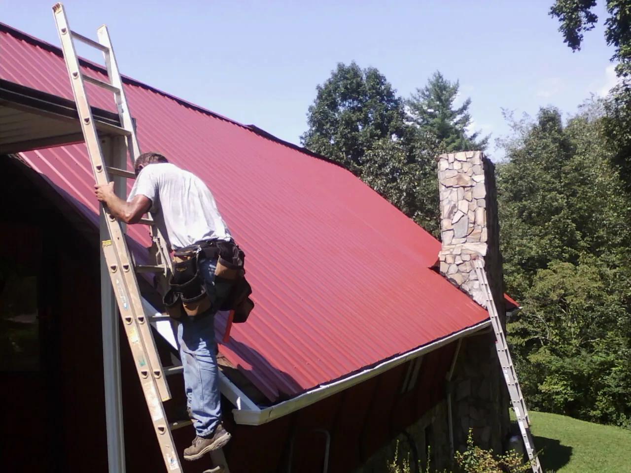 Roofer on a ladder beside a red metal roof and stone chimney — Soffit & Fascia Repair by Roofing Brothers in Belgrade